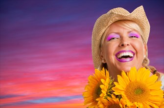 Beautiful laughing girl wearing cowboy hat holding yellow sunflowers against purple and pink sunset