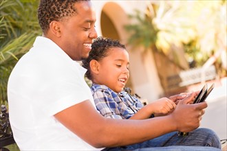 African american father and mixed-race son using computer tablet on bench in park