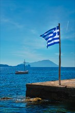 Greek flag in the blue sky on pier and traditional greek fishing boat in the Aegean sea with greek flag