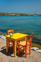Yellow cafe restaurant table of street cafe with chairs on beach in Adamantas town on Milos island with Aegean sea with boats and yachts in background. Milos island