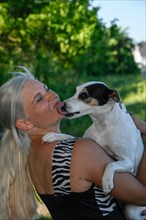 Im Garten steht jungen blonde huebsche Frau wird vom Hund im Arm mit ausgestreckter Zunge gekuesst