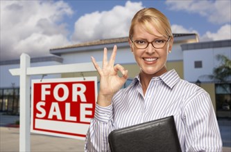 Smiling businesswoman with okay sign in front of vacant office building and for sale real estate sign