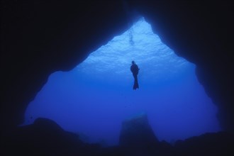 Diver floating in open water in front of large cave entrance of underwater cave