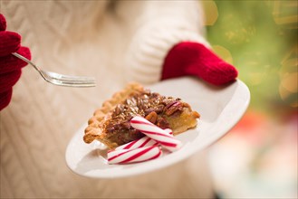 Woman wearing red mittens holding plate of pecan pie with peppermint candy against decorated tree and lights
