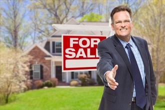 Smiling male agent reaching for a hand shake in front of beautiful house and for sale real estate sign