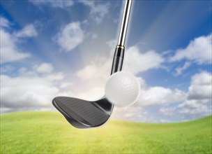 Black golf club wedge iron hitting golf ball against grass and blue sky background