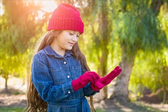 Cute mixed-race young girl wearing red knit cap putting on mittens outdoors