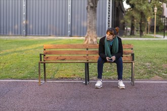 Latin woman sitting on a bench in a park with her cell phone in her hands looking to the side pensively