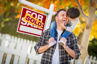 Young mixed-race chinese and caucasian father and son in front of sold for sale real estate sign and fall yard