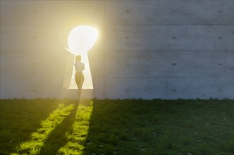 Woman standing at bright sun light shining through keyhole in concrete wall in grass and rocks field