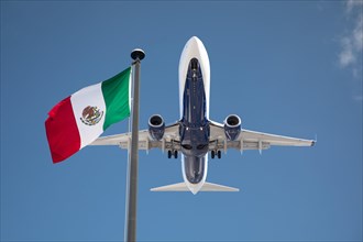Bottom view of passenger airplane flying over waving Mexico flag on pole