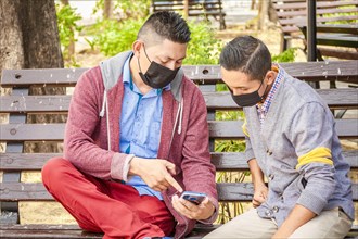 Two men wearing masks checking their cell phones on a bench