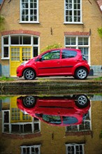 Red car on canal embankment in street of Delft with reflection