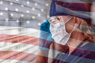 Stressed female doctor or nurse on break at window wearing medical face mask and goggles with ghosted american flag