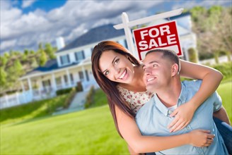 Playful excited military couple in front of home with for sale real estate sign