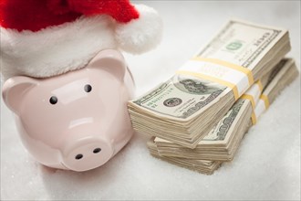 Piggy bank wearing santa hat near stacks of hundred dollar bills on snowflakes