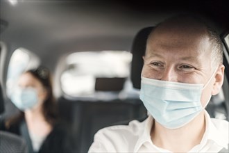 A happy taxi driver wearing a mask and the passenger on the back seat of the car in the background