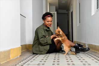 Im Flur sitzt eine junge huebsche Afro-Amerikanische Frau und spielt mit ihrem kleinen braunen Hund. Die schwarzen Stiefel mit den Netzstruempfen und der gruenen Jacke ergeben einen laessigen und frec...