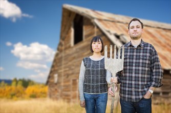 Biracial chinese and caucasian young couple holding wooden pitch fork in front of rustic barn in the country