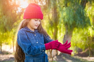 Cute mixed-race young girl wearing red knit cap putting on mittens outdoors