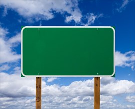 Blank green road sign with wooden posts over blue sky and clouds