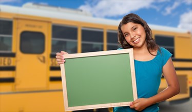 Young female hispanic student with blank chalkboard near school bus