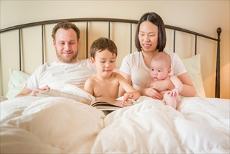 Young mixed-race chinese and caucasian baby boys reading a book in bed with their father and mother