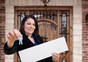 Hispanic woman holding blank sign and keys in front of house