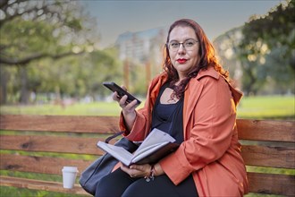 Woman sitting on a bench in a park checking her agenda and cell phone while having a coffee