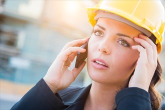 Young worried looking professional female contractor wearing hard hat at construction site using cell phone