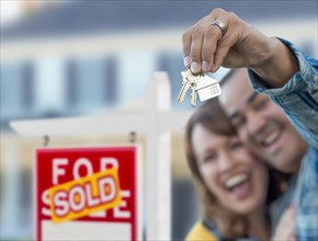 mixed-race couple in front of sold real estate sign and house with keys