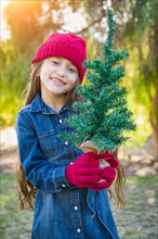 Cute mixed-race young girl wearing red knit cap and mittens holding small christmas tree outdoors