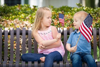 Young sister and brother comparing each others american flag size on the bench at the park