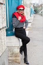 In einem Park lehnt an einer Mauer eine gut aussehende Dame mit roter Muetze und passendem Schal. Die schwarze Hose mit den Stiefeln passt zum Pullover im Matrosenlook. Die grosse schwarze Sonnenbrill...