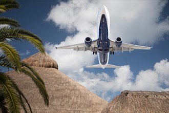 Bottom view of passenger airplane flying over tropical palm trees and huts