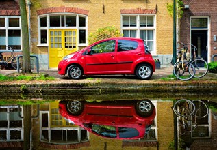 Red car on canal embankment in street of Delft with reflection and bicycle