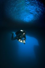 Diver dives with underwater lamp through rock grotto underwater cave Blue Dome with blue light in Blue Hole of Gozo