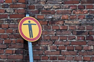 Round traffic sign with man gesturing with outstretched arms in front of brick wall