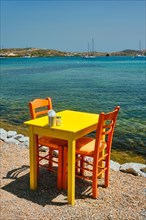 Yellow cafe restaurant table of street cafe with chairs on beach in Adamantas town on Milos island with Aegean sea with boats and yachts in background
