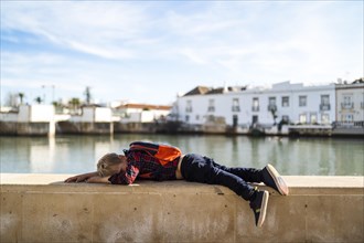 Tired preschooler or kid tourist with backpack resting on the riverbank in Tavira