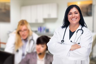 Handsome hispanic doctor or nurse standing in her office with staff working behind