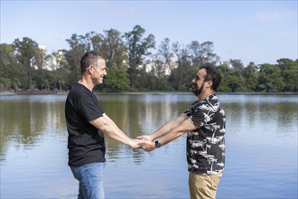 Homosexual mature white male couple holding hands smiling at a lake