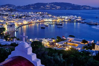 Mykonos Chora town Greek tourist holiday vacation destination with famous windmills port with boats yachts illuminated in evening blue hour with St. Basil church cross. Mykonos