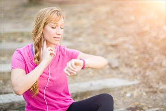Young fit adult woman outdoors in workout clothes listening to music with earphones checking her heart rate
