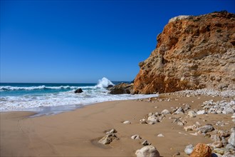 Portugal hat an der Algarve viele Steilkuesten mit feinem Sandstrand und hohen Wellen des Atlantik. Der blaue Himmel mit der weissen Gischt ist ein guter Kontrast zu den roetlichen Felsen