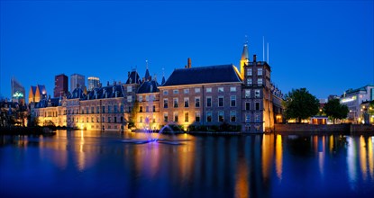 View of the Binnenhof House of Parliament and the Hofvijver lake with downtown skyscrapers in background illuminated in the evening. The Hague