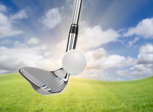 Chrome golf club wedge iron hitting golf ball against grass and blue sky background