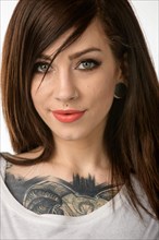 Portrait im Fotostudio einer jungen huebschen Frau langen braunen Haaren und einem Tattoo auf der Brust. Sie blickt direkt zur Kamera