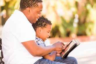 African american father and mixed-race son using computer tablet on bench in park