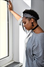 Eine nachdenkliche Afro-Amerikanische junge huebsche Frau steht an einem Fenster und blickt nachdenklich zu Boden. Sie traegt einen grauen Pullover mit einem passenden Stirnband